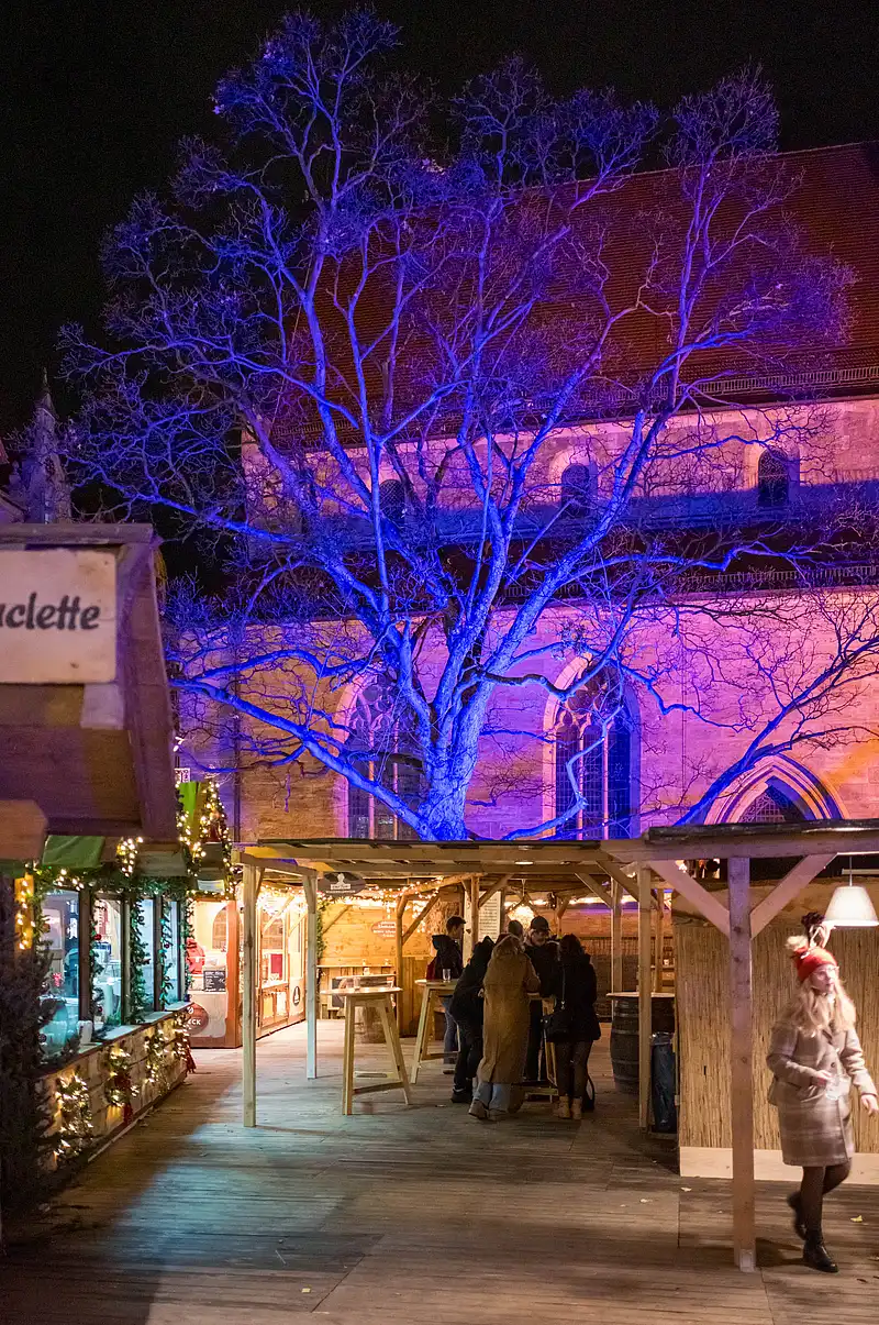 Belebte Marktstraße bei Nacht mit blauem Lichterbaum, warmen Lampen an Buden und einer Frau auf dem Weg