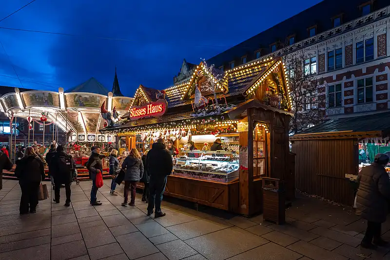Weihnachtlicher Anger bei Nacht: mehrere Stände, der „Süßes Haus“-Schriftzug und warmes Licht vor vollem Platz.