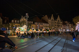Weihnachtsmarkt am Platz bei Nacht: farbig beleuchtete Stände, wartende Besucher und Tramschienen im Vordergrund