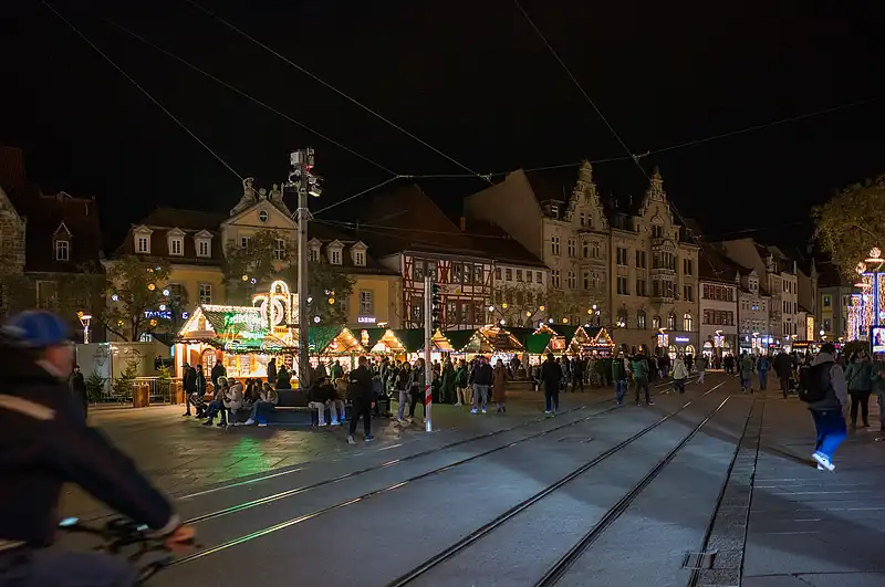 Weihnachtsmarkt am Platz bei Nacht: farbig beleuchtete Stände, wartende Besucher und Tramschienen im Vordergrund