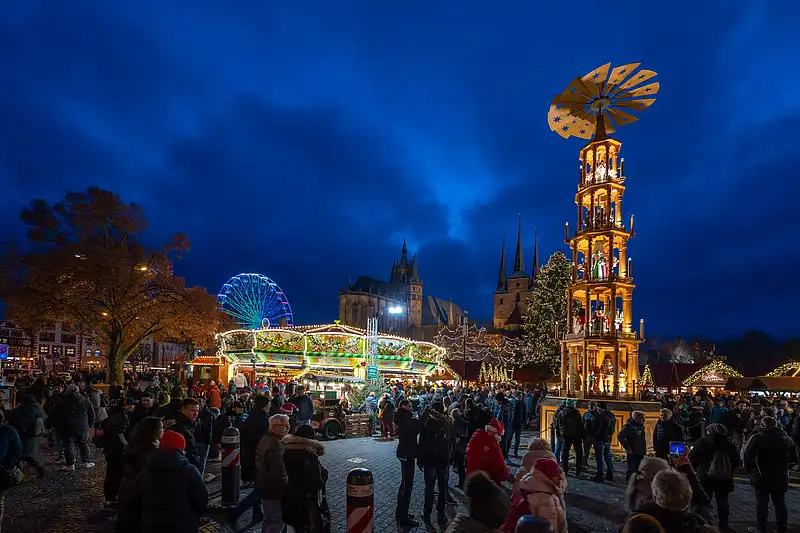 Blick über den Weihnachtsmarkt mit leuchtender Pyramide, geschmücktem Baum und voller Menge vor der Domkulisse.