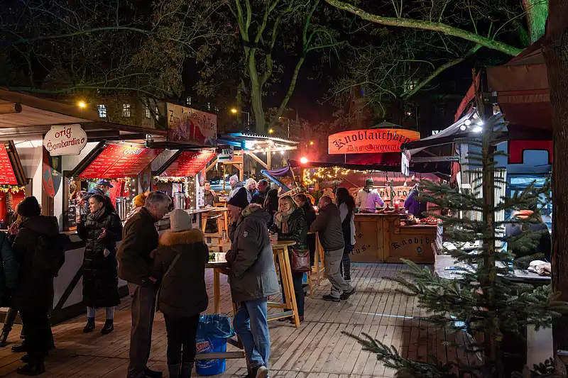 Viele Gäste an leuchtenden Ständen am Abend, warmes Licht in den Buden und festliche Stimmung draußen.