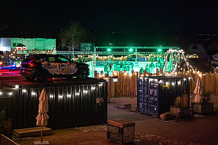 Grün beleuchteter Eventbereich mit Absperrung und Containern; auf einem Container steht ein Auto als Blickfang.