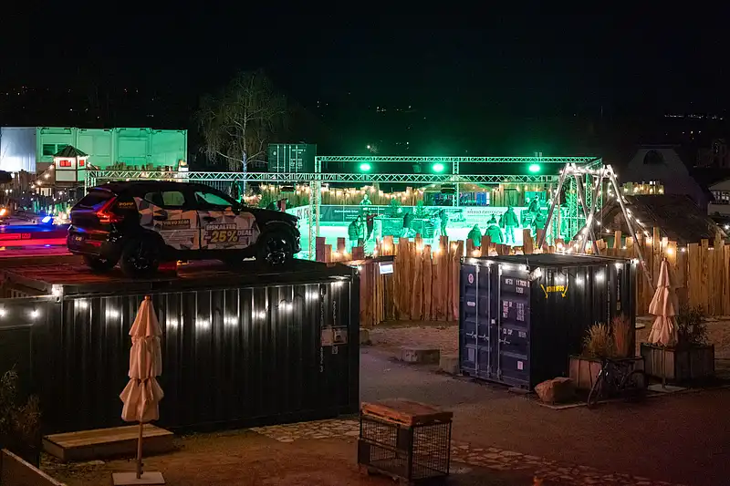 Grün beleuchteter Eventbereich mit Absperrung und Containern; auf einem Container steht ein Auto als Blickfang.