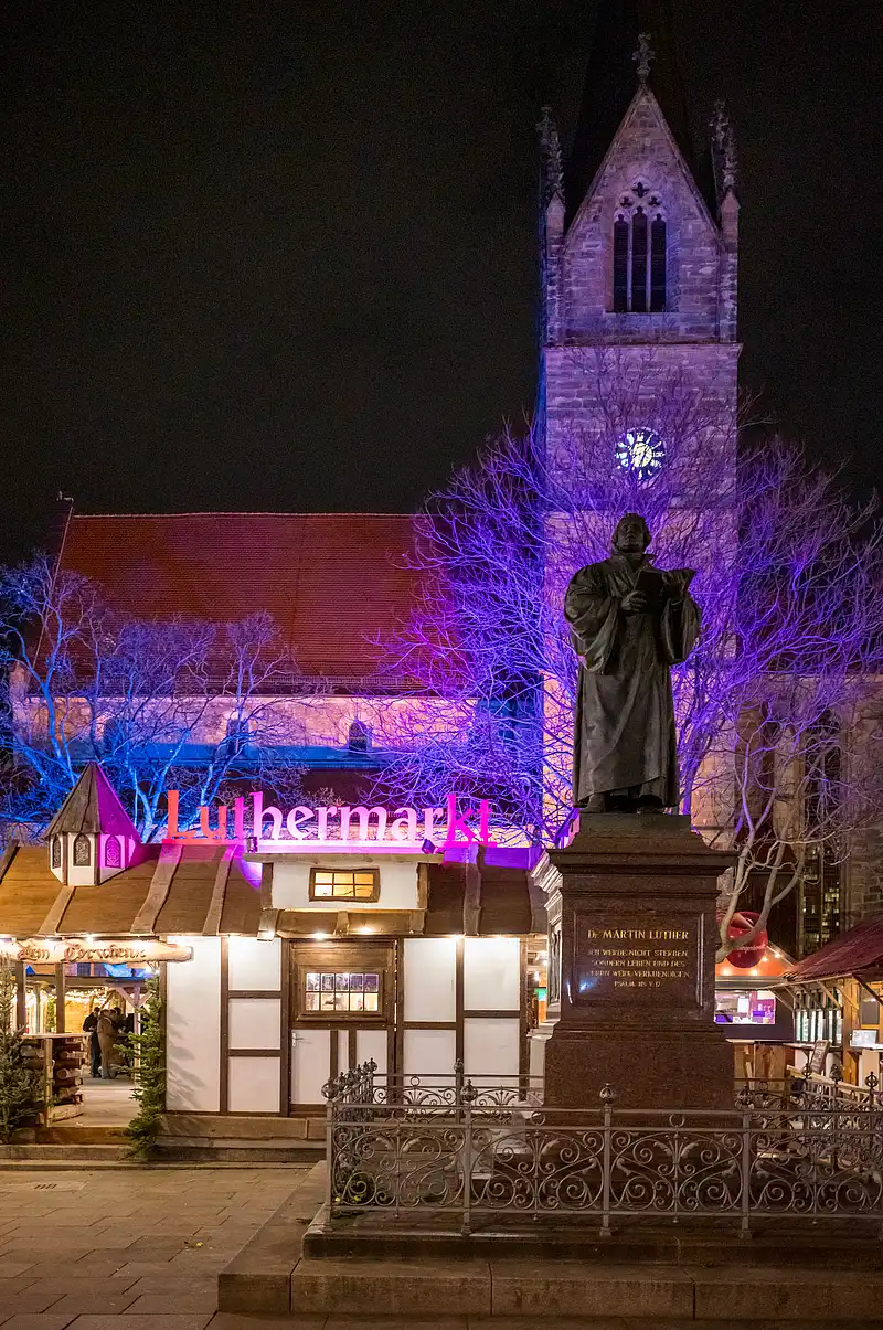 Luthermarkt mit Luther-Denkmal im Fokus, dahinter Turm der Kaufmannskirche im Violettlicht und beleuchtete Holzbuden am Abend