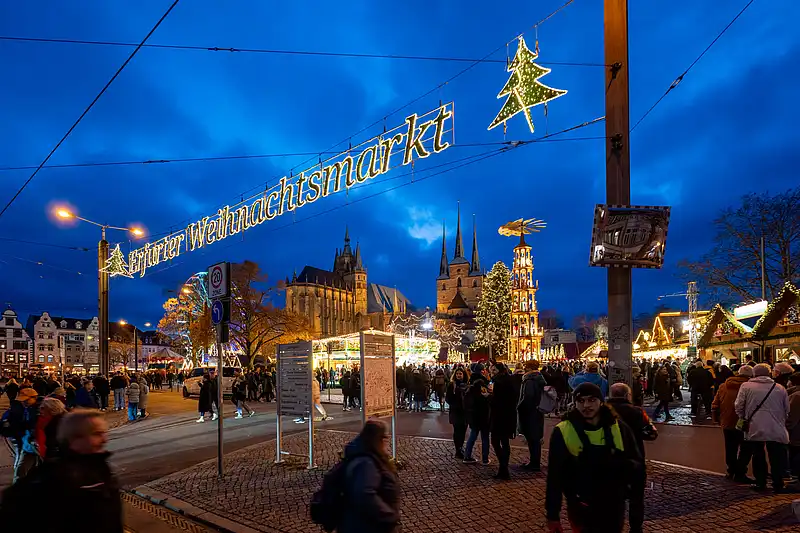 Seitliche Perspektive auf den leuchtenden Schriftzug; davor viele Besucher, rechts starten Budenreihen am Platzrand.