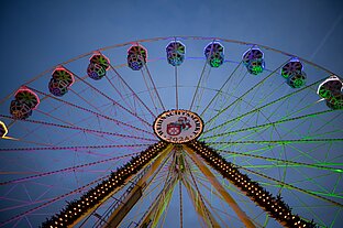 Blick von unten auf ein Riesenrad mit farbig beleuchteten Gondeln vor tiefblauem Abendhimmel.
