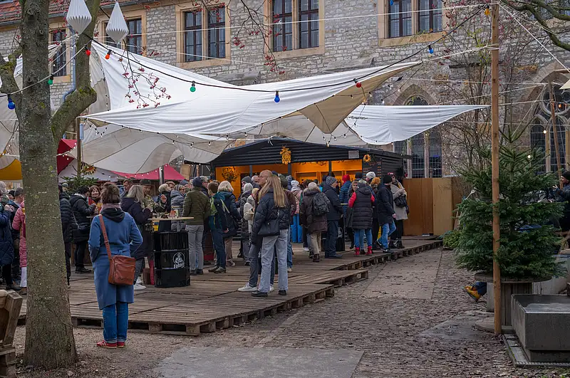 Gedrängter Weihnachtsmarkt im Innenhof: Besucher unter weißen Marktzelten, festliche Lichterketten und bunte Deko am Weg