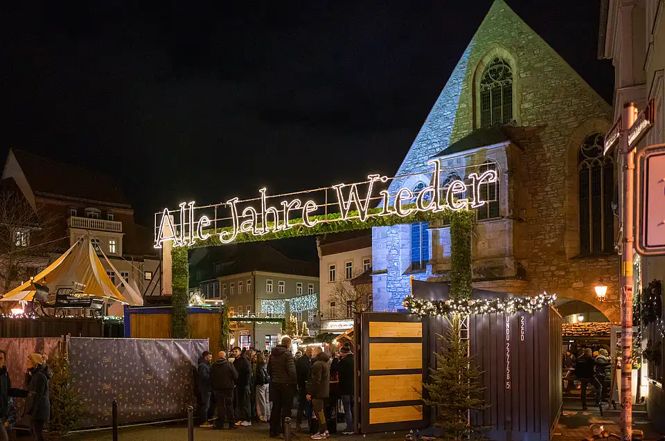 LED-Schriftzug „Alle Jahre wieder“ über dem Markt, beleuchtete Kirche im Abendlicht und Besucher an Ständen.