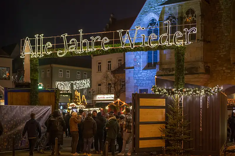 Menschenmenge vor blau angestrahlter Kirche, darüber leuchtet Alle Jahre wieder, Tannen im Vordergrund