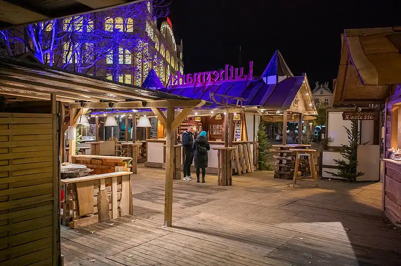Langer Durchgang zwischen beleuchteten Marktständen, blau-violette Fassadenlichter und zwei Spaziergänger auf dem Platz