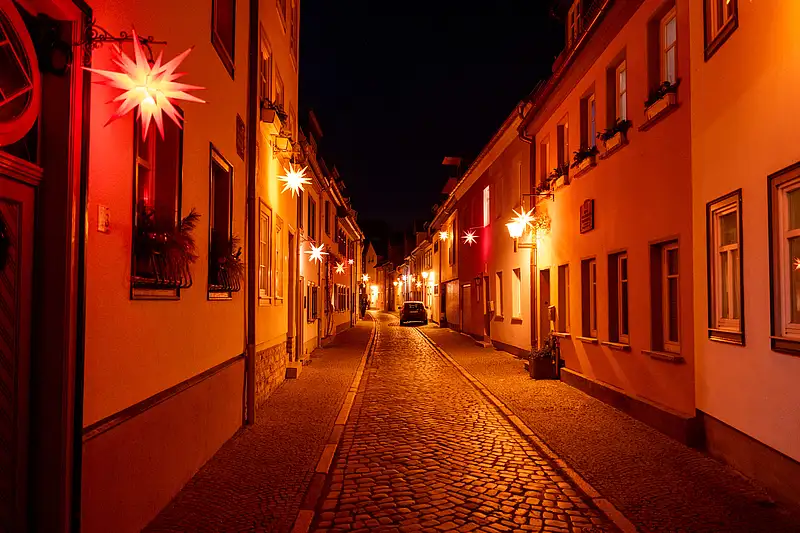 Weihnachtsmarkt-Straße bei Nacht: rote Beleuchtung, Sterne an Fassaden und Passanten auf gepflastertem Weg