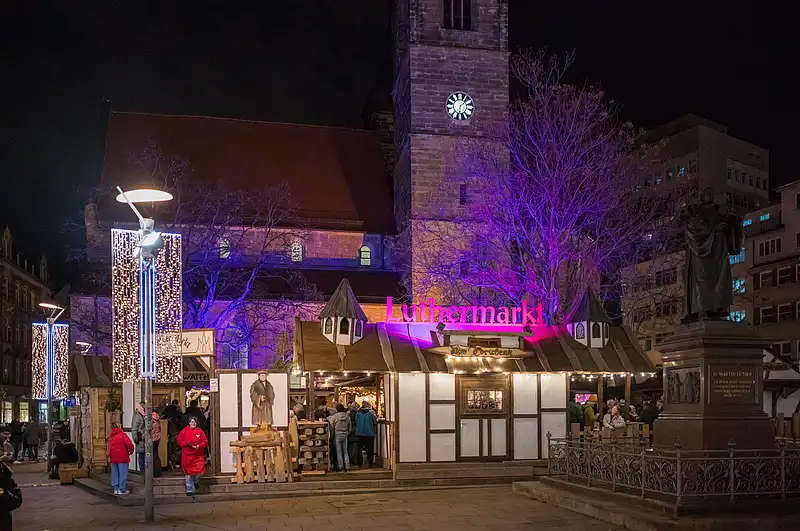 Eingang zum Luthermarkt mit leuchtendem Schriftzug, dahinter Kirchturm und violett angestrahlte Bäume.
