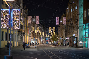 Weihnachtlich beleuchteter Anger bei Nacht, Lichterketten über den Gleisen, wenige Passanten auf dem Pflaster.