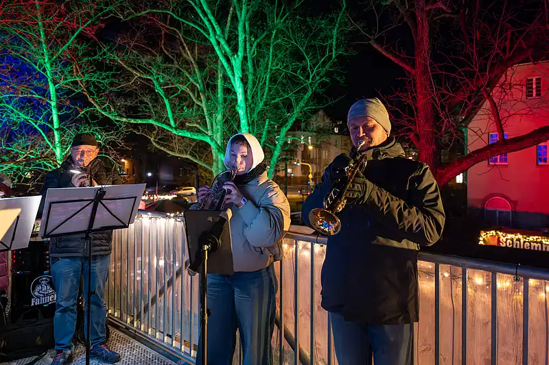 Bläserensemble musiziert am Weihnachtsmarkt, Sängerin im Kapuzenmantel vor grün-roten Lichtbäumen und Holzgeländer.