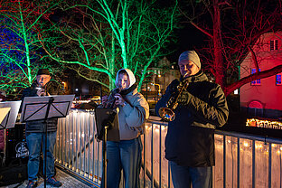 Bläserensemble musiziert am Weihnachtsmarkt, Sängerin im Kapuzenmantel vor grün-roten Lichtbäumen und Holzgeländer.