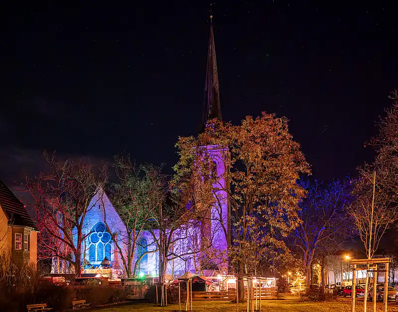 Illuminierte Bäume im Winter: violette und orangefarbene Lichtakzente vor beleuchteter Kirchenfront und Marktständen.