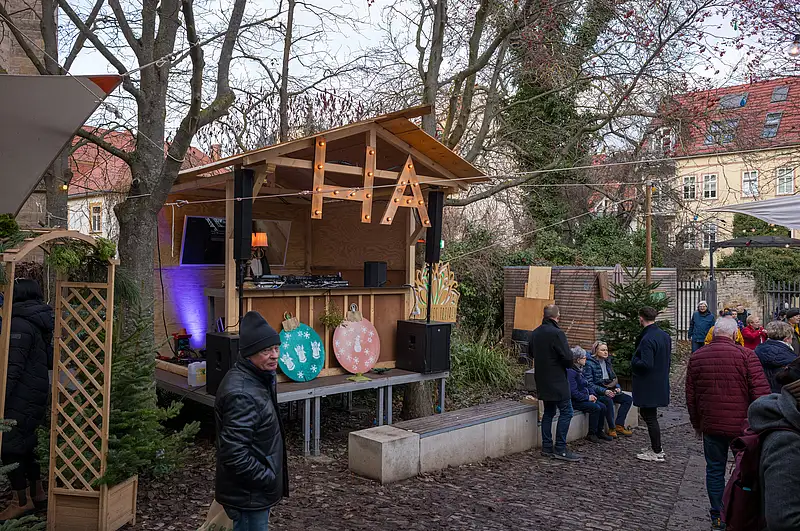 Weihnachtsmarktstand im Garten: beleuchtete Holzbar mit farbigen Ornamenten und wartenden Besuchern am Abend