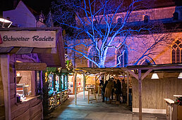 Schild "Schweizer Raclette" an der Holzfront, blau beleuchteter Baum und Besucher auf dem Weg durch die Gasse