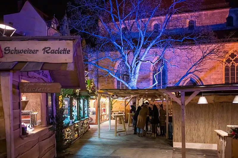 Schild "Schweizer Raclette" an der Holzfront, blau beleuchteter Baum und Besucher auf dem Weg durch die Gasse