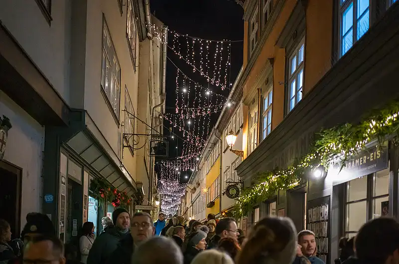 Weihnachtsmarkt in der Gasse: dicht hängende Lichterketten, Passanten flanieren vorbei an Eissalon und grüner Dekoration