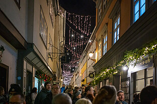 Weihnachtsmarkt in der Gasse: dicht hängende Lichterketten, Passanten flanieren vorbei an Eissalon und grüner Dekoration