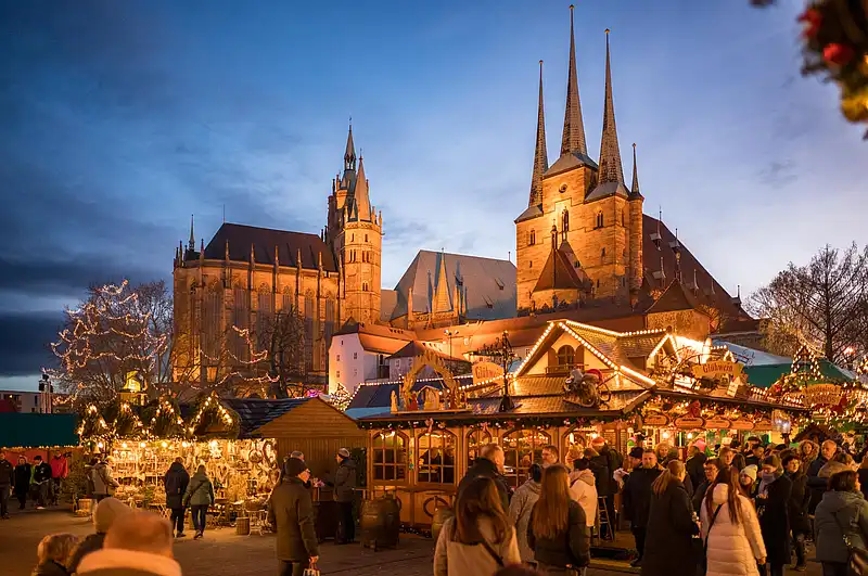 Besucher zwischen Glühweinstand und Lichterketten, im Hintergrund leuchten Dom und hohe Kirchtürme am Abend.
