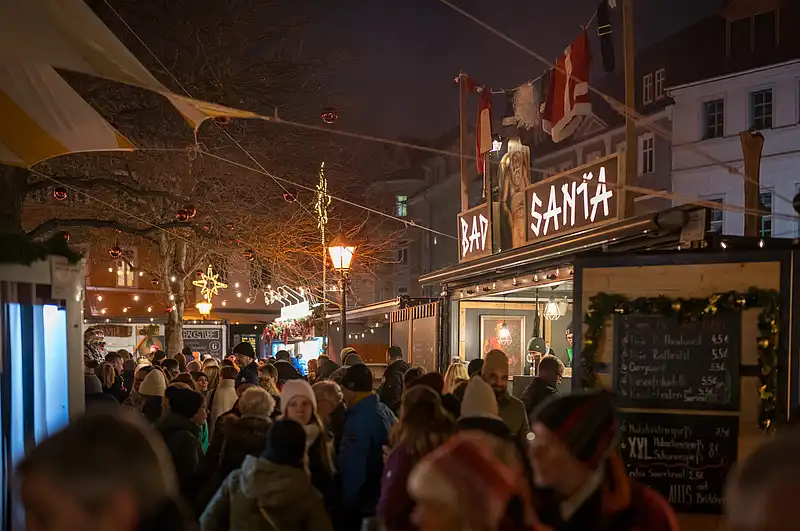 Besucher stehen in beleuchteter Marktgasse, vorn ein Stand mit Menütafel und warmen Lichterketten