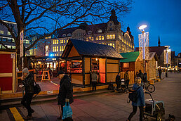 Am blauen Abend leuchten Holzbuden am Platz, Passanten gehen vorbei und im Hintergrund funkeln Lichtvorhänge