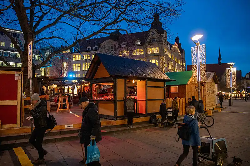 Am blauen Abend leuchten Holzbuden am Platz, Passanten gehen vorbei und im Hintergrund funkeln Lichtvorhänge
