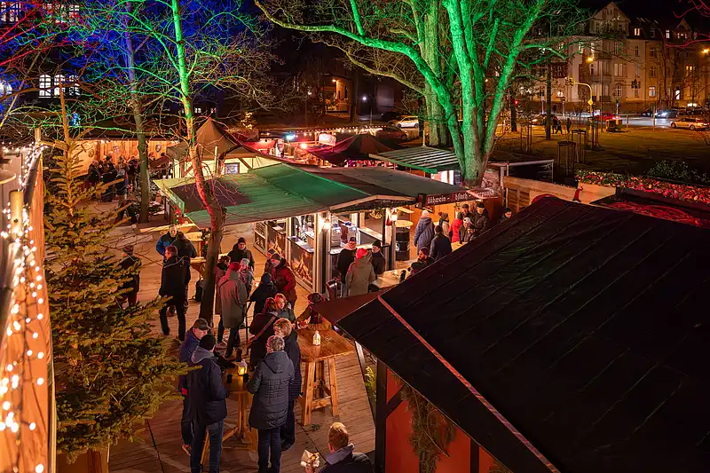 Überblick über den Weihnachtsmarktweg bei Nacht: beleuchtete Stände, Besuchergruppen und Bäume entlang der Gasse.