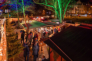 Überblick über den Weihnachtsmarktweg bei Nacht: beleuchtete Stände, Besuchergruppen und Bäume entlang der Gasse.