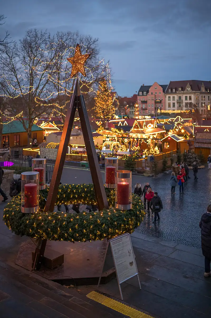Großer Adventskranz mit leuchtendem Stern und Kerzen, dahinter Budenlichter und Besucher in der Dämmerung.