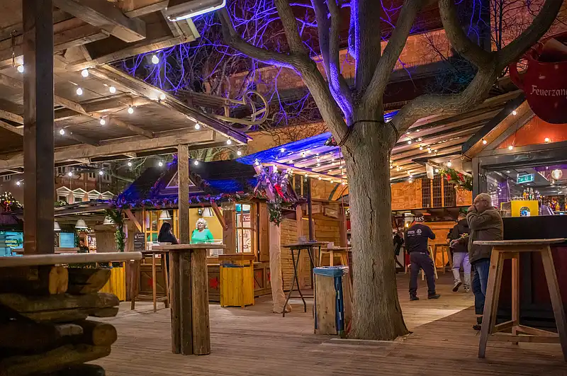 Ruhige Marktlaube am Luthermarkt: Holzbänke, warme Lampen und ein großer Baum mit blauem Licht im Vordergrund