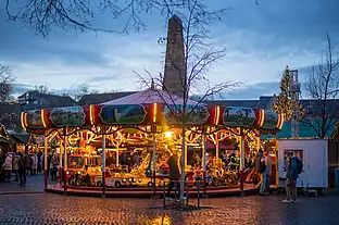 Beleuchtetes Kinderkarussell mit kleinen Autos, Besucher auf Kopfsteinpflaster, Abendstimmung am Domplatz.