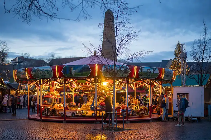 Beleuchtetes Kinderkarussell mit kleinen Autos, Besucher auf Kopfsteinpflaster, Abendstimmung am Domplatz.