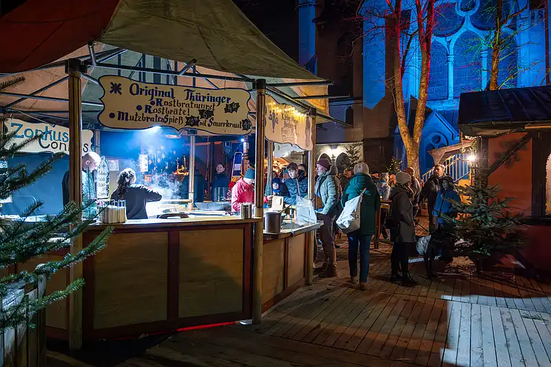 Rostbratwurst-Stand am Markt: Menschen schlendern am Rauch vorbei, während blau angestrahlte Fassaden den Weg säumen.