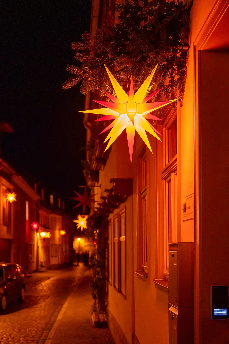 Weihnacht bei Nacht: großer leuchtender Stern vor Fichten und beleuchteter Fassadenzeile in der Glockenquergasse