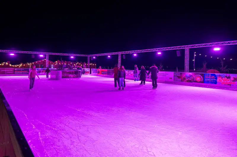 Weitwinkel auf die Eislaufbahn: Schlittschuhläufer auf großer Eisfläche, violette Spots und dunkler Nachthimmel.