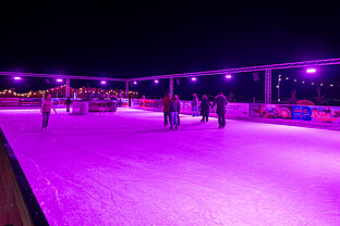 Weitwinkel auf die Eislaufbahn: Schlittschuhläufer auf großer Eisfläche, violette Spots und dunkler Nachthimmel.