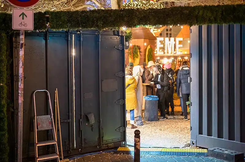 Blick durch das geöffnete Tor in den Weihnachtsmarkt: Besucher warten am Stand, Lichter und Tannen