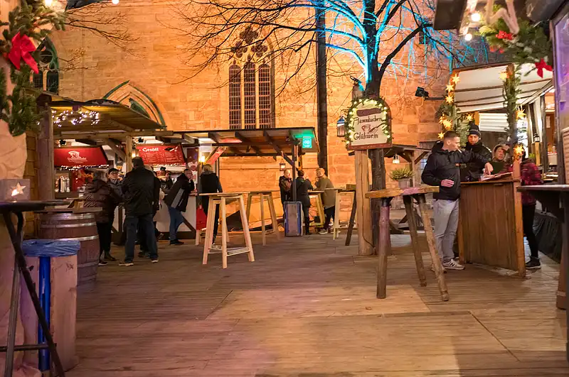 Weihnachtsmarkt-Gasse mit Holzständen, beleuchtetem Baum und Besuchern an der Theke vor der Kaufmannskirche