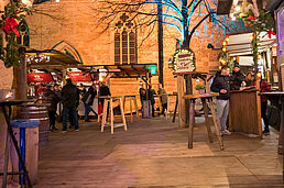 Weihnachtsmarkt-Gasse mit Holzständen, beleuchtetem Baum und Besuchern an der Theke vor der Kaufmannskirche