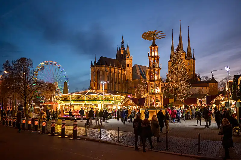 Panoramablick auf Riesenrad, Karussell und Weihnachtspyramide, davor flanieren Menschen über den Platz bei Dämmerung.