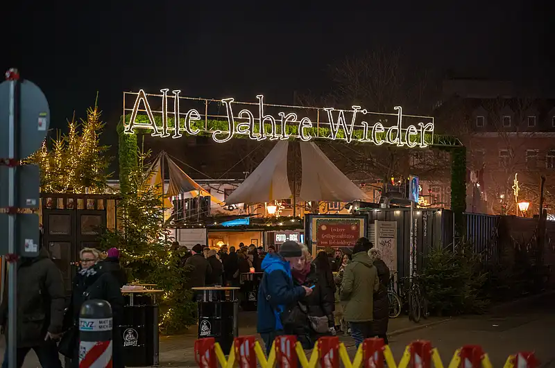 Belebter Weihnachtsmarkt-Eingang bei Nacht mit leuchtendem Schriftzug, Tannen und warmen Lampen vor Buden