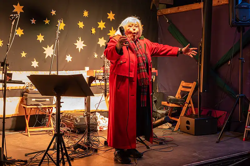 Gerda Gabriel im roten Mantel auf Weihnachtsmarkt-Bühne, Sternchenmotiv im Hintergrund und warmes Licht.