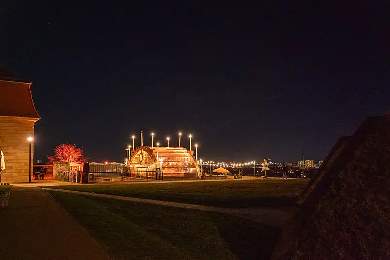 Weitblick bei Nacht: beleuchteter Pavillon des Weihnachtsmarkts auf dem Hügel, Lichterkette und Stadt im Hintergrund.