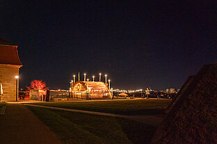 Weitblick bei Nacht: beleuchteter Pavillon des Weihnachtsmarkts auf dem Hügel, Lichterkette und Stadt im Hintergrund.