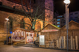 Holzstände am Platz mit großem Werbebild, Fahrrad vor einer Tafel und warm beleuchteter Baumkrone in der Nacht