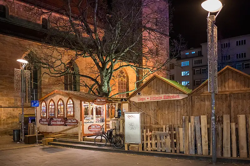Holzstände am Platz mit großem Werbebild, Fahrrad vor einer Tafel und warm beleuchteter Baumkrone in der Nacht